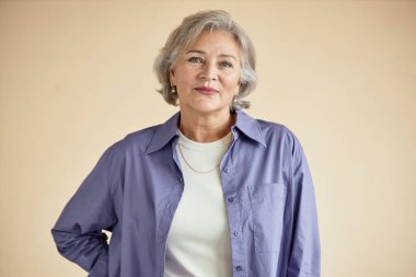 Beyaz bluz ve lavanta gömlek giyerken gülümseyen yaşlı beyaz bir kadının portresi. Sakin duruşu ve dostça ifadesiyle tarafsız bir arkaplanda duruyor.