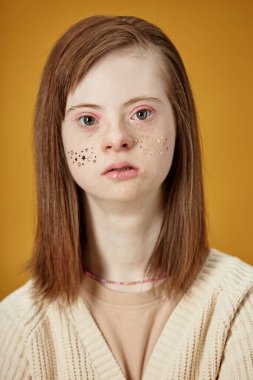 Portrait of young woman with Down syndrome facing camera, having freckles and long straight hair worn loose against mustard-yellow backdrop, presenting calm expression clipart