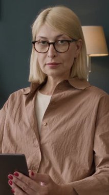 Kameraya poz verirken dijital tablet kullanan kendinden emin beyaz kadın psikoloğun dikey orta boy portresi.