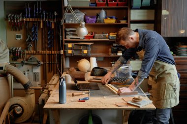 İyi organize edilmiş bir atölyede çalışan yetenekli bir zanaatkar, kesin ölçümlere odaklanıyor. Arka planda görünen çeşitli araçlar ve bileşenler üretken bir atmosfer yaratıyor.