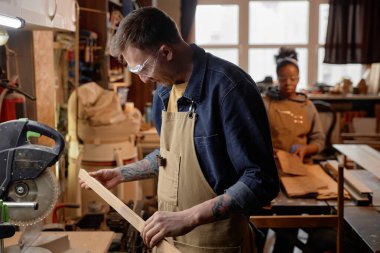 Zanaatkar atölyesinde çalışan bir adam, etrafındaki aletlere ve tahta parçalarına odaklanmış. Arka plandaki kişi de kırsal ahşap atölyesinde zanaat faaliyetinde bulunuyor.