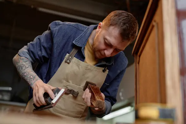 stock image Woodworker concentrating on sanding wooden furniture piece, wearing apron and surrounded by workshop tools and equipment. Tattoed arms visible while engaged in meticulous craftsmanship