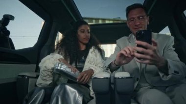 Lüks arabaların arka koltuğunda sosyal medya için çirkin giysiler giymiş, akıllı telefonlardan fotoğraf çeken ve pencerelerden resimlerini çeken iki ünlü çiftin orta boy fotoğrafı.
