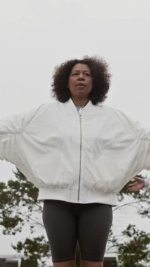 Orta yaşlı Afrikalı Amerikalı bir kadının dikey olarak yoga minderinin üzerinde dikey ve yavaş çekimleri. Gri gökyüzünün altında sabah sporu yaparken derin bir nefes alıyor.