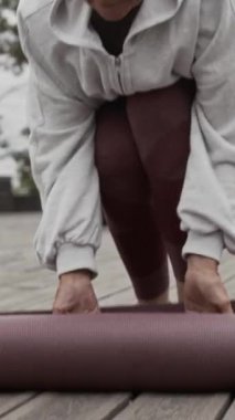 Aktif giyimli yaşlı beyaz kadının Piblic Park 'taki ahşap zemine eğitim için yoga minderi sererken dikey yavaş çekimleri.