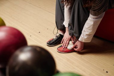Bowling salonundaki maça hazırlanırken bovling ayakkabılarını bağlamak. Ahşap zemin ve arka planda görülen renkli bowling topları canlı bir atmosfer yaratıyor.