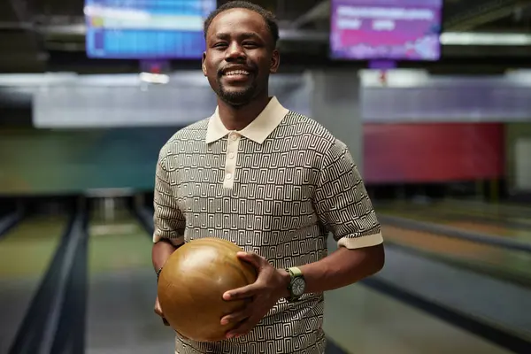 stock image African American man holding bowling ball while standing at bowling alley, smiling warmly, wearing patterned shirt and wristwatch, bowling lanes visible in background