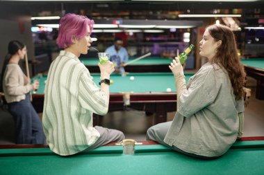 Group of friends enjoying conversation and drinks while playing billiards at a dimly lit lounge. People engaging in leisure activity and bonding over a shared hobby clipart