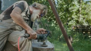 Beyaz erkek esnaf yavaş yavaş doğal taşları parlatmak için yonga makinesi kullanırken bahçede heykel yapıyor.