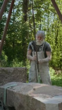 Beyaz tenli üst düzey bir adamın metal zincirleri çekerken bahçedeki ağır taşları kaldırdığı dikey bir fotoğraf.