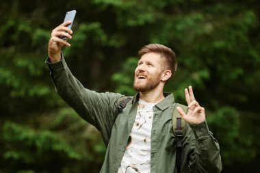 Sakallı adam elini uzatmış telefonu tutarken gülümsüyor, yemyeşil ormanın ortasında neşeli bir ifade sergiliyor.