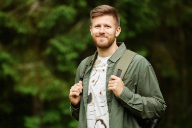 Beyaz bir adamın portresi, ormanda sırt çantası takmış ve kayışlarını tutarak gülümseyen ve doğrudan kameraya bakarak doğanın özünü yakalayan bir adam.