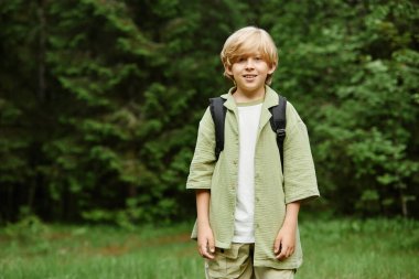 Genç beyaz çocuk ormanda duruyor, sırt çantası taşıyor ve hafifçe gülümsüyor. Arkaplan yemyeşil ağaçlar ve otlarla dolu maceraperest ruhu ve dingin doğayı yakalıyor.