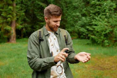 Yeşil bir çevrede elinde sprey şişesiyle dikilen bir adam günlük kıyafetlerini giyerken işine odaklanıyor ve etrafı çimen ve ağaçlarla çevrili.