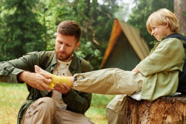 Baba, oğullarını bir ağaç kütüğünde otururken bacağını bandajlıyor. Kamp gezisinde bakım ve doğa aktivitelerine odaklanıyor.