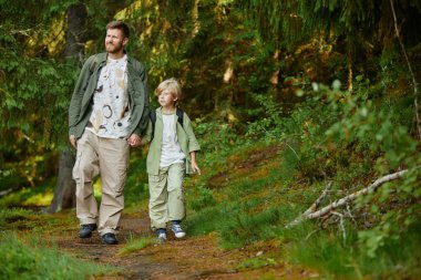 Baba ve oğul, güneşli bir günde yeşil orman yolunda el ele yürüyorlar. İkisi de günlük yürüyüş kıyafetleri giyip doğanın ve birlikteliğin tadını çıkarıyor.
