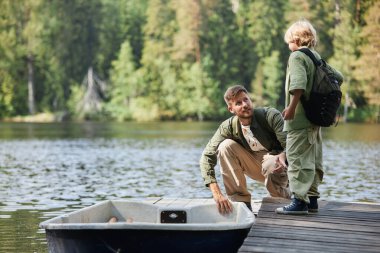 Babam rıhtımda diz çökmüş gölün kenarında oğluyla konuşurken ağaçlarla çevrili, ikisi de balıkçı kıyafeti giymiş.