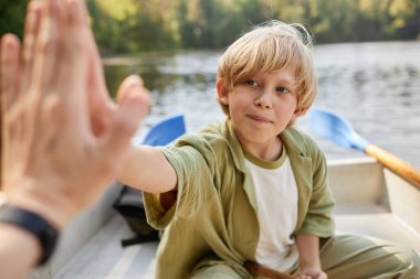 Sarı saçlı genç bir çocuk göldeki bir kanoda oturuyor ve birine beşlik çakıyor. Arka planda ağaçlar ve suyla huzurlu bir manzara var.