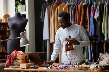 İkinci el dükkanında sosyal ağların hesabını tutarken dizüstü bilgisayar kullanarak klasik fotoğraf makinesini satışa sunan şık tişörtlü bir erkek Afro-Amerikan ikinci el mağaza asistanı.