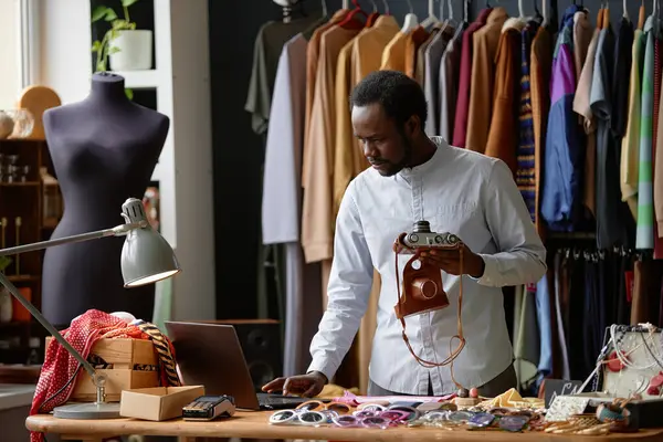 Stock image Male African American thrift store assistant in classy shirt using laptop putting up vintage photo camera for sale while keeping account in social networks at second hand shop, copy space