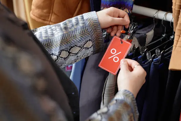 Hands of female shopper choosing second hand clothes with red discount tag offered for sale at thrift store, copy space