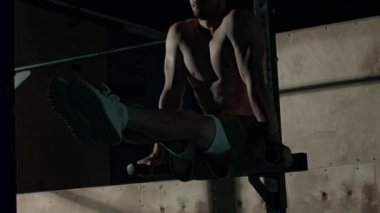 Atletik genç Kafkasyalı üstsüz adam spor salonunda karanlıkta antrenman yaparken paralel barda bacak bükme hareketi yapıyor.
