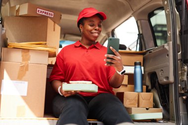 Gülümseyen teslimat işçisi elinde paketle teslimat aracındaki akıllı telefona bakarken etrafı gülen ve kırmızı üniforma giyen kutularla çevrili.