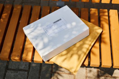 Posta paketi ve zarf, dış taraftaki ahşap bankta, yaprakların arasından süzülen güneş ışığı tarafından kısmen gölgeleniyor.