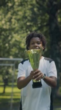 Genç Afrikalı Amerikalı erkek futbolcunun şampiyonluğu kazandıktan sonra altın kupayı kaldırdığı, güneşli bir günde sahada durup kameraya gülümsediği dikey yavaş bir çekim.