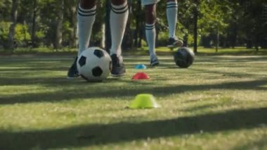 Spor üniforması giyen iki Afrikalı Amerikalı genç futbolcunun çimenlik alanda erkek koçla antrenman yaparken külahlar arasında futbol topu sallamaları.