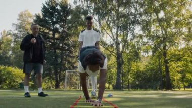 İki Afrikalı Amerikalı erkek futbol takımının açık hava eğitimi sırasında çimenlikte çeviklik testi yaparken beyaz yetişkin koçun onları izlediği tam kare.