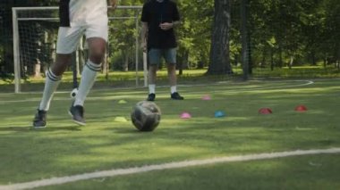 Siyah-beyaz futbol forması giyen Afro-Amerikalı genç çocuk, beyaz erkek antrenör ile antrenman sırasında çimenli alanda top sürüyor.