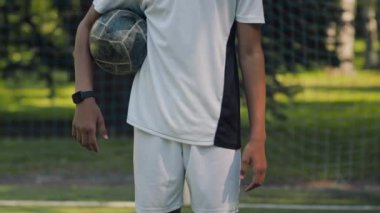 Siyah-beyaz üniformalı Afro-Amerikan genç futbolcunun orta kesiminde çimenli sahada topu kolunun altına alarak kameraya poz veriyor.