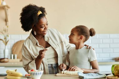 Orta halli mutlu Afrika kökenli Amerikalı anne ve küçük kız muzlu smoothie yaparken mutfak tezgahında birlikte kahvaltı yaparken bej duvarlara yaslanarak,