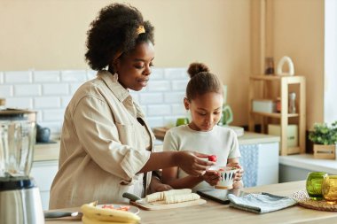 Orta boy Afrika kökenli Amerikalı anne ve küçük kızı kahvaltıda bej mutfakta sağlıklı meyve suyu için çilek tartıyorlar.