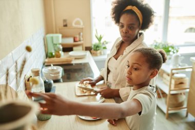 Afro-amerikan küçük bir kızın birlikte sağlıklı yemek pişirdiği ve annesinin bej renkli güneşli mutfakta meyveli kahvaltı malzemelerini almak için uzanıp uzaya ulaştığı yan manzara.