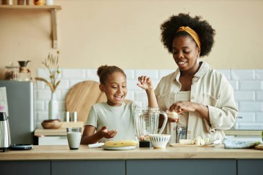 Gülümseyen Afrikalı Amerikalı kız ve genç anne büyük tezgahta muzlu smoothie yapıyor. Sıcak bej mutfakta eğleniyor ve mutlu anları paylaşıyorlar.