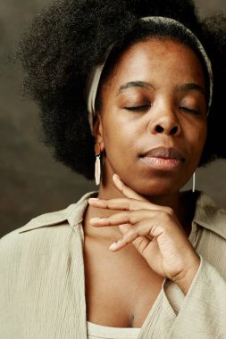 Gözleri kapalı, makyajsız, zarif bir şekilde yüzüne dokunan, stüdyodaki kahverengi arka planda tedaviden sonra pürüzsüz cildin tadını çıkaran, şehvetli, genç Afrikalı Amerikalı kadının portresini yakından çek.
