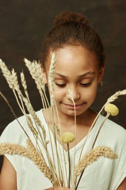Beyaz elbiseli, elinde bir buket kurumuş çiçekle stüdyodaki kahverengi arka plana bakan Afro-Amerikan genç kızın yakın çekimi.