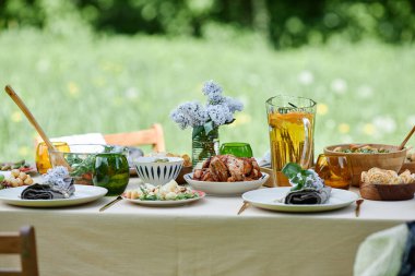 Çeşitli yemekler, taze çiçekler ve ferahlatıcı içeceklerden oluşan açık hava yemek masası seti. Güzel yeşil arka plan sakin ve davetkar bir atmosfer yaratır.