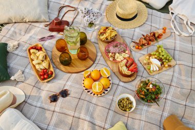 Kareli battaniyede sergilenen yiyecek, meyve, salata, abur cubur ve içeceklerle birlikte piknik aksesuarları arka planda açık hava yemeklerinin özünü yansıtıyor.