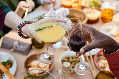 Çeşitli yemekler ve mutlu atmosferle dolu neşeli akşam yemeği etkinliği sırasında şarap ve kokteyl kadehleri kaldıran melez ırktan insanların yakın çekimi.