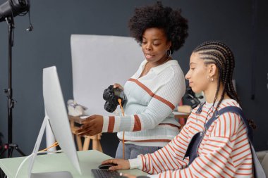 İki Afro-Amerikalı genç kadının, fotoğraf stüdyosunda çalışan ve bilgisayar ekranındaki resimlere bakan kadın fotoğrafçı portresi.