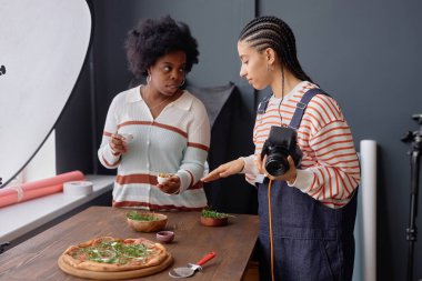 Portrait of two Black young women as team of female photographers collaborating during food photography set and staging fresh rustic pizza on wooden table clipart