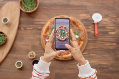 Mobil yemek fotoğrafçılık seti sırasında akıllı telefon tutan ve ahşap masada lezzetli pizzaların fotoğrafını çeken kadınların üst görüntüsü.