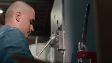 Kafası tıraşlı, mavi azot eldiveni ve kot ceketli, üretim tesisinde çalışırken makina teçhizatına basan, konsantre beyaz erkek işçilerden oluşan bir sandık.