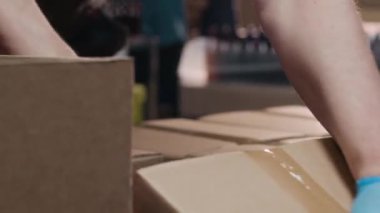 Tanımlanamayan sanayi işçisinin mavi nitril eldivenli, karton kutuları elle bantla paketlediği yakın plan çekimleri.