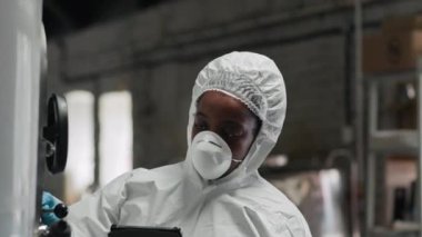 Elma şarabı fabrikasındaki depodaki paslanmaz çelik tankı incelerken dijital tablet üzerine notlar alan tam korumalı takım elbiseli siyahi kadının belini kaldırın.