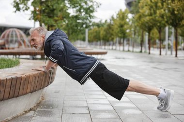Spor giyimli, formda, kenarda şınav çeken ve şehir parkında fayanslarla çalışan yaşlı bir adamın yan görüntüsü.