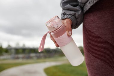 Bulutlu bir havada şehir parkında koşarken su sızdırmaz kapağı olan pembe plastik su şişesini tutan kadın eline yaklaş, boşluğu kopyala